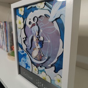 Puede incluir: Una obra de arte de papel recortado enmarcada que presenta un personaje del videojuego Genshin Impact. El personaje, Ganyu, est&aacute; representado con un atuendo azul y blanco sobre un fondo blanco y azul. La obra est&aacute; enmarcada en un marco blanco con un passe-partout negro.