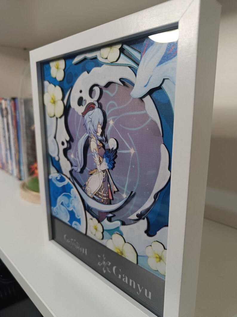 Puede incluir: Una obra de arte de papel recortado enmarcada que presenta un personaje del videojuego Genshin Impact. El personaje, Ganyu, est&aacute; representado con un atuendo azul y blanco sobre un fondo blanco y azul. La obra est&aacute; enmarcada en un marco blanco con un passe-partout negro.