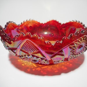 Puede incluir: Un cuenco de vidrio de carnaval rojo vibrante con un acabado iridiscente y un borde festoneado. La superficie exterior muestra un patrón prensado detallado que consiste en formas geométricas, motivos de estrellas y un diseño de rejilla de diamantes.
