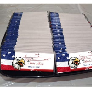 May include: A tray of folded white napkins with a patriotic design. The napkins feature a red, white, and blue color scheme with an eagle graphic and text that reads "THE COURTS FOR Mark Mone May 10, 2025".