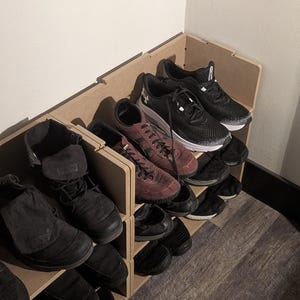 Puede incluir: Un zapatero de madera lleno de varios zapatos. Los zapatos son de diferentes estilos y colores, incluyendo zapatillas negras, botas marrones y zapatos de vestir negros. El zapatero es de madera de color marrón claro.