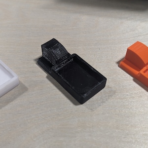 K&ouml;nnte beinhalten: Drei 3D-gedruckte Kunststoffschalen in Wei&szlig;, Schwarz und Orange. Jede Schale hat eine kleine, erh&ouml;hte Lippe an einer Seite und eine rechteckige Vertiefung in der Mitte.