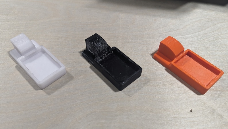 K&ouml;nnte beinhalten: Drei 3D-gedruckte Kunststoffschalen in Wei&szlig;, Schwarz und Orange. Jede Schale hat eine kleine, erh&ouml;hte Lippe an einer Seite und eine rechteckige Vertiefung in der Mitte.