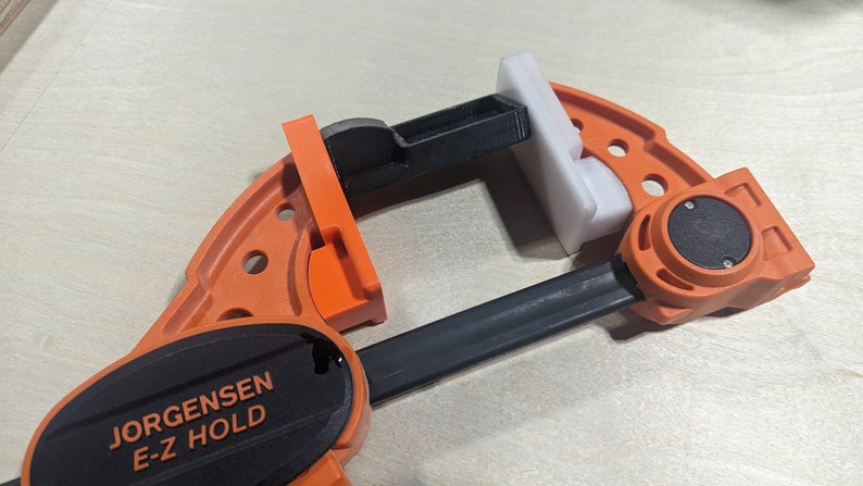 K&ouml;nnte beinhalten: Eine orangefarbene und schwarze Jorgensen E-Z Hold Zwinge mit einem wei&szlig;en Kunststoffteil in den Backen. Die Zwinge ist f&uuml;r die Holzbearbeitung konzipiert und hat einen schwarzen Griff.