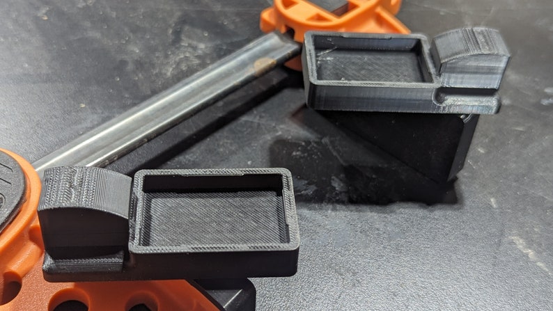 K&ouml;nnte beinhalten: Zwei schwarze 3D-gedruckte Eckklemmen mit einer rechteckigen &Ouml;ffnung. Die Klemmen sind f&uuml;r die Verwendung mit einer Holzbearbeitungsklemme konzipiert.