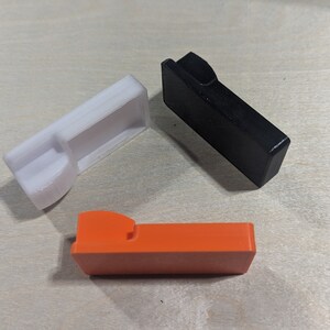 K&ouml;nnte beinhalten: Drei Plastikteile in verschiedenen Farben: Wei&szlig;, Schwarz und Orange. Jedes Teil hat eine einzigartige Form mit einem Ausschnitt.