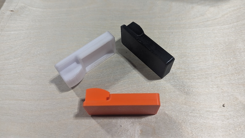 K&ouml;nnte beinhalten: Drei Plastikteile in verschiedenen Farben: Wei&szlig;, Schwarz und Orange. Jedes Teil hat eine einzigartige Form mit einem Ausschnitt.