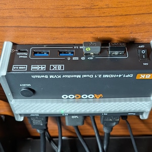 May include: A black and grey 8K DP1.4+HDMI 2.1 Dual Monitor KVM Switch by AOOCOO. The switch has two HDMI ports, two DP ports, a USB 3.0 port, and a wired remote port. It also has a select button and an on/off switch.