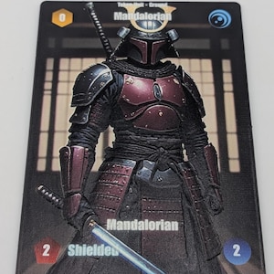 Puede incluir: Una tarjeta coleccionable rectangular que muestra un personaje mandaloriano con armadura de color rojo oscuro y negro, casco y katana. La tarjeta incluye el texto Mandalorian, Token Unit - Ground, Shielded y los números 0, 2 y 2 en las esquinas.