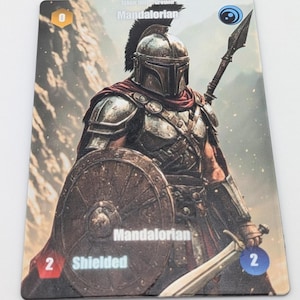 May include: A rectangular trading card featuring a warrior in metallic armor and a helmet with a black crest. The figure holds a round wooden shield and a spear. Text on the card includes Mandalorian, Shielded, and numerical values 0 and 2.