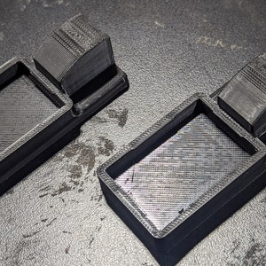 K&ouml;nnte beinhalten: Zwei schwarze 3D-gedruckte Kunststoffschalen mit einem erh&ouml;hten rechteckigen Abschnitt auf einer Seite. Die Schalen sind so konzipiert, dass sie kleine Gegenst&auml;nde aufnehmen k&ouml;nnen.