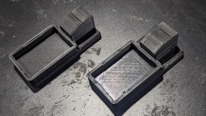 K&ouml;nnte beinhalten: Zwei schwarze 3D-gedruckte Kunststoffschalen mit einem erh&ouml;hten rechteckigen Abschnitt auf einer Seite. Die Schalen sind so konzipiert, dass sie kleine Gegenst&auml;nde aufnehmen k&ouml;nnen.