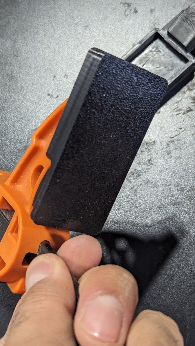 K&ouml;nnte beinhalten: Eine schwarze Kunststoffklemme mit einer strukturierten Oberfl&auml;che. Die Klemme ist an einer orangefarbenen Kunststoffbasis befestigt.