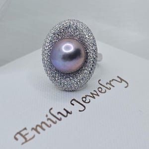 May include: A silver ring with a large, round, iridescent pearl set in a halo of sparkling white stones. The ring is on a white box with the text "Emily Jewelry" written in cursive.