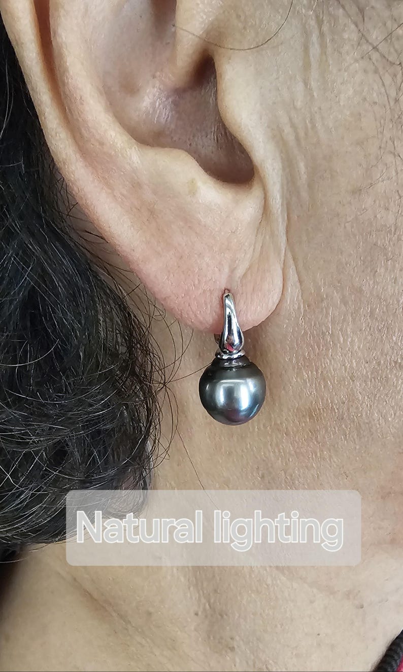 May include: A pair of silver hoop earrings with a dark gray pearl dangling from each hoop. The earrings are worn by a person with dark hair. The image is labeled "Natural lighting".