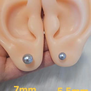 May include: Two gray pearl stud earrings, 7mm and 5.5mm, displayed on a flesh-toned ear model. The earrings are round and have a subtle sheen. The background is a soft, blurred white.