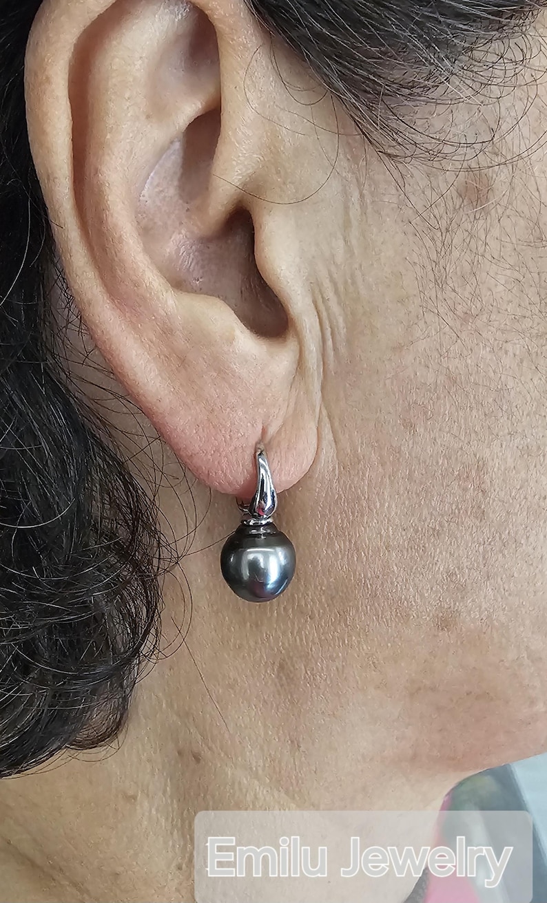 May include: A pair of silver hoop earrings with a single black pearl dangling from each hoop. The earrings are worn by a person with dark hair.