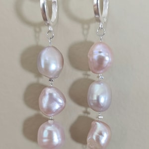 以下が含まれることがあります： 銀製のフープピアス。各フープから3つのピンク色の淡水パールがぶら下がっている。