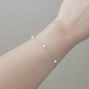 May include: A delicate silver chain bracelet with five small white pearls. The bracelet is worn on a person's wrist.