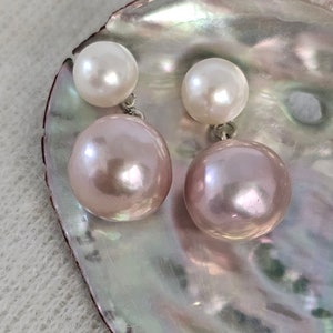 May include: A pair of pearl earrings. Each earring features a small, white pearl stud and a larger, pinkish-purple pearl drop. The earrings are displayed on a pearlescent, iridescent surface, creating a soft, elegant aesthetic.