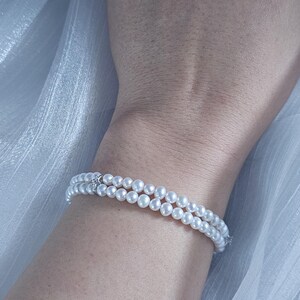 May include: A double-strand pearl bracelet with a small silver clasp. The bracelet is worn on a wrist, with a soft, white fabric in the background. The pearls are round and white, and the bracelet appears delicate.