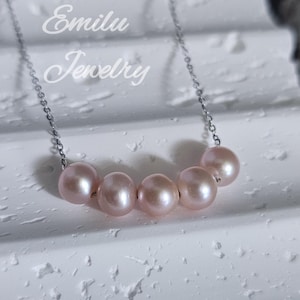 以下が含まれることがあります： シルバーチェーンに5つのピンクパールをあしらった繊細なネックレス。 ジュエリーは、テクスチャ加工された白い表面に表示されています。 背景には「Emilu Jewelry」の文字が見えます。