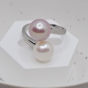 May include: A bypass ring featuring a polished silver-tone band with two round pearls. One pearl is a soft lavender-pink hue, while the other is a classic creamy white. The pearls are positioned at opposite ends of the open, curved metal band.