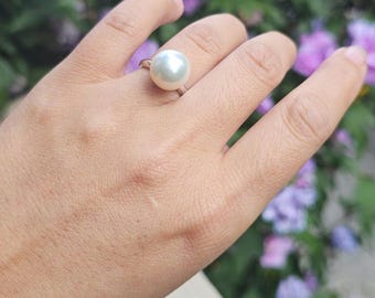May include: A silver ring with a large, round, white pearl. The ring is on a hand, with a blurred background of purple flowers and green foliage. The text "Natural lighting" is at the bottom.
