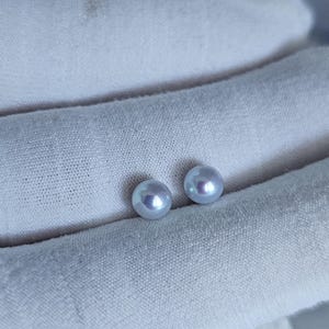 May include: Two small, round, iridescent pearls rest on a white, textured fabric. The pearls have a subtle, light blue hue and a smooth, polished surface. The fabric appears to be a soft, woven material, creating a delicate contrast with the pearls.