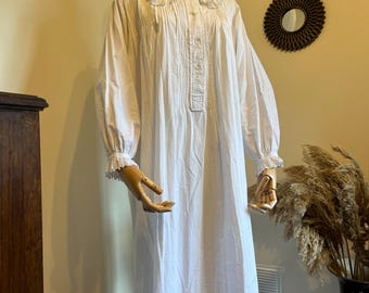 May include: A long, white nightgown on a mannequin. The nightgown features a ruffled collar, button-down front, and long sleeves with ruffled cuffs. The fabric appears to be lightweight cotton or linen. The garment is floor-length.