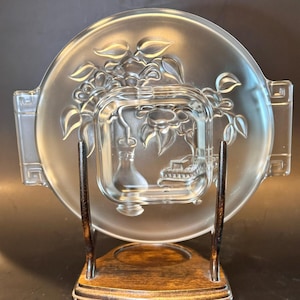 May include: A clear glass serving dish with a floral design, featuring a central square bowl and a round outer plate with handles. The dish is displayed on a wooden stand with a dark finish, showcasing its elegant design.