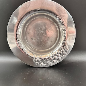 May include: A silver-colored plate with a raised center and a decorative grape and leaf design around the rim. The plate has a shiny, reflective surface and appears to be made of metal.