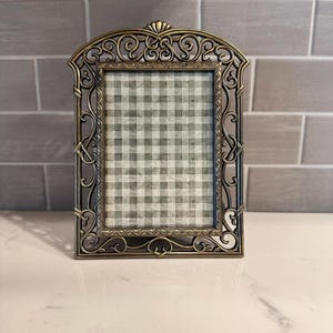 May include: Ornate bronze-colored picture frame with a decorative scroll design. The frame has a rectangular opening with a gray and white checkered pattern visible inside. The frame is made of metal and has an arched top.