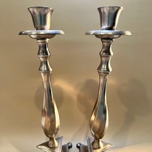Puede incluir: Un par de candelabros plateados. Cada candelabro tiene una parte superior acampanada, un plato redondo y un tallo retorcido y alargado. La base es cuadrada con pies redondeados. Los candelabros se muestran sobre un fondo neutro.