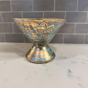 May include: A decorative, gold-colored bowl with a flared top and a pedestal base. The bowl features a textured design with blue accents. The interior and exterior are painted with gold and blue tones, creating a unique, artistic look.