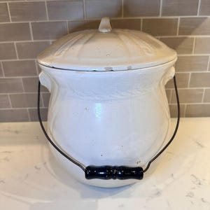 May include: A white ceramic crock with a lid and a black handle. The crock has a rounded shape with a lid that has a small handle. The handle is black with a dark blue accent. The crock is sitting on a white surface.