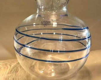 May include: Clear glass vase with a round body and a flared neck, adorned with horizontal blue stripes. The rim is trimmed with blue. The vase has a label that reads "DANSK International Designs China".