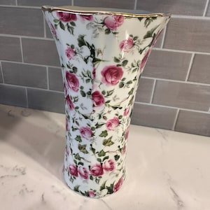 May include: A white ceramic vase featuring a repeating pattern of pink roses and green ivy leaves. The vase has a flared, scalloped rim with a thin gold-painted edge. The body of the vase is cylindrical with a slight taper toward the base.