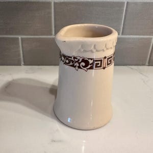 May include: Cream-colored ceramic creamer with a decorative brown band featuring floral and geometric patterns. The creamer has a scalloped rim and a curved, flared base. A vintage-style kitchenware item.