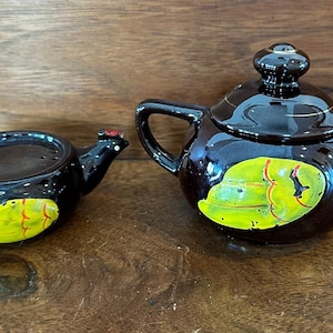 May include: A set of two brown ceramic teapots with a yellow and red rooster design. The smaller teapot has a rooster head on the spout and the larger teapot has a rooster head on the handle.