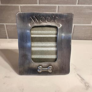 May include: A rectangular brushed silver metal picture frame with a vertical orientation. The word "WOOF" is embossed at the top center, and a raised bone-shaped icon is centered at the bottom. The frame contains a striped gray and white insert.