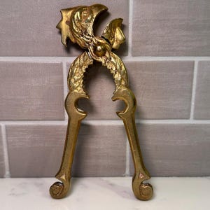 May include: A gold-colored metal nutcracker featuring an ornate, textured design on the handles and a stylized bird-like head at the top. The tool has two long, curved handles that meet at a central pivot point, with a serrated inner grip area.