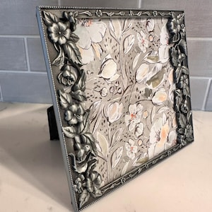 May include: A square picture frame with a metallic silver finish featuring a raised, three-dimensional floral and leaf relief pattern along the borders. The frame contains a print with a light gray background and white floral illustrations.