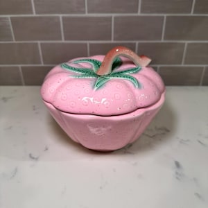 May include: A pink ceramic strawberry-shaped trinket box with a lid. The lid features green leaves and a red-orange stem. The box has a textured surface and is sitting on a white surface.
