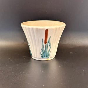 May include: A small, off-white ceramic bowl with a flared shape and vertical ribbed texture. The bowl is decorated with a hand-painted design of cattails in shades of brown, green, and blue. The bowl is likely used for serving or display.