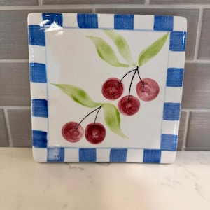 May include: Square ceramic serving dish with a white center featuring a cherry branch design with red cherries and green leaves. The border is blue and white checkered. The dish is on a white surface.