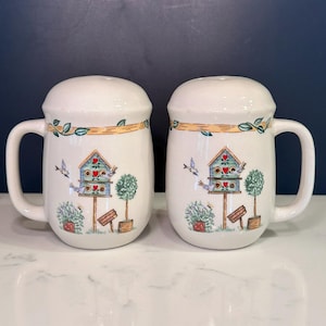 May include: A pair of off-white ceramic salt and pepper shakers, each shaped like a mug with a handle. The surface features a painted design of a blue birdhouse with red hearts, small birds, a green topiary tree, and a horizontal branch border.