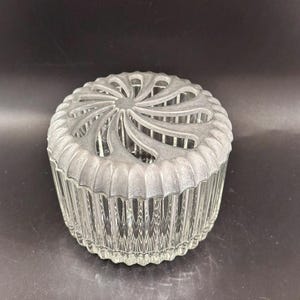 May include: A clear glass jar with a ribbed design and a decorative silver-colored lid. The lid features a spiral pattern with open spaces. The jar is round and sits on a dark surface.