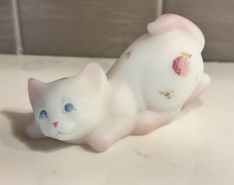Puede incluir: Una figura de cristal esmerilado blanco de un gato en una postura de estiramiento juguetona. El gato presenta ojos azules pintados a mano, una nariz rosa y pequeños detalles florales en rosa y amarillo en la espalda y la cabeza, con un tono rosado.