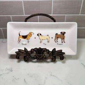 May include: A rectangular white ceramic tray featuring a graphic illustration of a Beagle plus a Pug equals a Puggle. The tray is displayed on a dark bronze-colored metal stand with ornate, scroll-like detailing along the front base.